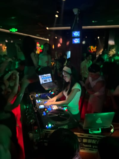 The image depicts a dimly lit, crowded scene, likely a nightclub or bar, with individuals engaged in various activities. The primary focus is on a woman in the foreground wearing a white top and a headscarf, appearing to be operating a DJ mixing console. Her hands are positioned over the controls, suggesting she is actively mixing music.
Several other people are visible in the background and surrounding areas, though their faces are mostly obscured or out of focus due to the low light and motion. Some individuals appear to be dancing or socializing, while others are partially visible behind the DJ. The overall atmosphere seems energetic, with the presence of a DJ and multiple people suggesting a lively event.
Prominent objects include the DJ mixing console with illuminated buttons and displays, a laptop placed on top of it, and what appear to be small screens or monitors to the left of the DJ. Green and red lighting illuminates the scene, contributing to a vibrant, club-like ambiance. A sign with Chinese characters and the word "Thank you" is visible in the upper right portion of the image.
The time of day is likely evening or night, consistent with a nightclub setting. No specific weather cues are discernible. The location context of "Liangjiang, China" suggests the setting is within that region. While specific emotions are difficult to gauge due to the image quality and lighting, the overall impression is one of entertainment and social gathering. A small, green, illuminated figure, possibly a decorative item or a toy, is visible in the darker lower-left section of the image.