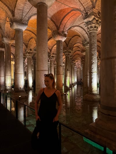 The image depicts the Basilica Cistern in Istanbul, Turkey. A woman in a black dress stands on a walkway in the foreground, looking down. The cistern is characterized by its extensive network of stone columns, supporting an arched brick ceiling. Water fills the floor of the cistern, reflecting the ambient lighting and creating a mirrored effect. Several other people are visible in the background, dispersed throughout the vast space. The atmosphere is dimly lit, with artificial lights highlighting the architectural features.