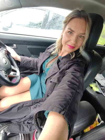 The image shows a blonde woman with bright pink lipstick sitting in the driver's seat of a car. She is wearing a teal-colored dress and a dark trench coat. Her right hand is on the steering wheel, and her legs are crossed. The car interior is visible, including the steering wheel, dashboard, and leather seats. The exterior of the car is blurred but appears to be an overcast day. The location is not specified in the image, and there are no visual cues to indicate it is Petropavlovsk-Kamchatsky, Russia.