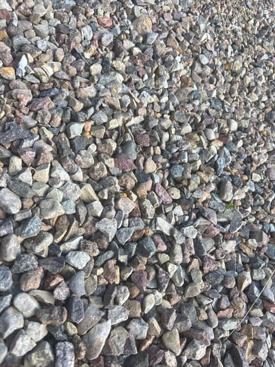 The image is a close-up, high-angle view of a pile of loose gravel. The gravel consists of small, angular stones in various shades of gray, white, pink, and brown. Some stones have speckles of darker colors. The gravel is densely packed, with individual stones overlapping and creating a textured surface. There are no people or other objects visible in the frame. The lighting suggests an outdoor environment, possibly during daylight. The image does not provide any specific information about its location in the city of Kapyĺ, Belarus.