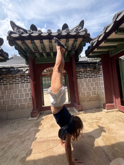 An athletic individual is captured in a handstand against the backdrop of a traditional Korean palace structure. The person, wearing a navy blue tank top and light grey shorts, is positioned with their legs extended vertically upwards. Their hands are planted firmly on the ground, supporting their entire body weight. The surrounding environment features traditional Korean architecture with ornate tiled roofs, intricate wooden structures painted in red and green, and stone walls. The ground is covered in a sandy, light-colored material. The sky above is blue with scattered white clouds. The scene appears to be a public space, possibly within a historical site in Seoul, South Korea, such as Gyeongbokgung Palace, given the architectural style.