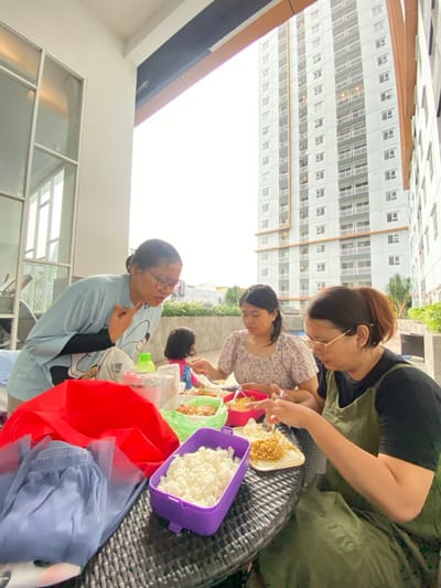 The image depicts an outdoor meal taking place on a paved terrace or balcony, identified as being in the city of Sidoarjo, Indonesia. Four individuals are visible, three adults and one child, gathered around a dark-colored, woven-texture outdoor table laden with food containers.
On the left, a woman with dark hair in glasses, wearing a light blue long-sleeved shirt, stands and leans forward, gesturing with her right hand (index and pinky fingers extended) and an open mouth, as if speaking or reacting. To her right, a second woman with dark hair, wearing a patterned short-sleeved top, is seated, looking down at a bowl of food and eating with a fork. Further to the right, a third woman with light brown hair in glasses, wearing a dark top and an olive-green apron-style dress, is seated, actively eating from a white plate with a fork. Partially visible behind the second woman is a young child in a pink top and blue bottoms, seated at the table. A hand from an unseen person is visible on the far right edge of the frame.
On the table, there is a purple rectangular container filled with white rice, a green container with what appears to be fried food, a pink bowl containing noodles, and a white plate with crispy noodles. Other items include a clear plastic container with a red lid, a green bottle, a red fabric item, and a light blue pleated skirt.
The immediate background behind the seated individuals features a low, light gray concrete wall, with some greenery beyond it. The wider background is dominated by a tall, modern multi-story apartment building, light in color with numerous balconies and windows, suggesting a residential urban environment. The sky overhead is bright white, indicating an overcast condition or overexposure. To the left, a structure with large windows and white frames is partially visible.