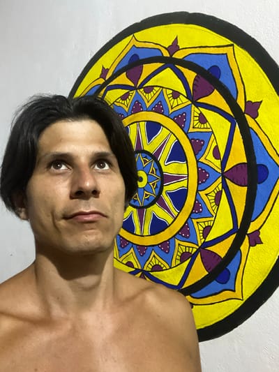 This is a medium shot, eye-level, outdoor photograph of a shirtless man with dark hair looking up and to his left, positioned in front of a large, colorful mandala painting on a white wall. The mandala features bold yellow, blue, and purple geometric patterns with black outlines. The man's face is in focus, showing his features and his gaze directed upwards. The background is mostly the white wall with the vibrant mandala on the right side. The lighting appears natural, illuminating the man's skin and the painting. The scene is set in Los Velásquez, Venezuela.