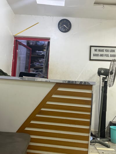 The image shows an indoor scene. On the left wall, there is a clock set to approximately 11:58, a sign that reads "WE MAKE YOU LOOK GOOD AND FEEL GOOD", and a doorway leading to what appears to be a storage or dressing area filled with clothing items hanging on racks. A yellow measuring tape is attached to the wall. To the right is a wooden staircase railing with vertical slats and a gray-colored object. A floor fan is visible towards the top of the image. The location is potentially Kubwa, Nigeria. The image contains no people.