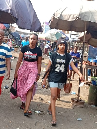 Three people stroll through a busy outdoor market. The woman on the left wears a black t-shirt with a red and white design, a long pink skirt, and carries a black purse. To her right, another woman in a black basketball jersey, light-colored shorts, and a beanie hat walks with a brown tote bag. The third person, only partially visible, wears a blue and white striped shirt and red pants. Overhead, several large umbrellas provide shade, while the market floor is littered with debris. Other people and stalls are visible in the background.