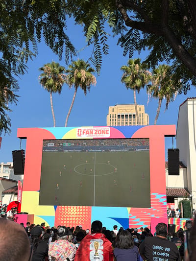 An outdoor public viewing of a soccer match is taking place on a large screen, likely at the Los Angeles World Cup Fan Zone in the city of Los Angeles, United States of America. The screen, which displays a soccer game in progress, is mounted on a vibrant, multi-colored structure. Spectators are seated and standing in front of the screen, facing it and appearing engaged with the game. The scene is set against a clear blue sky, with palm trees and buildings in the background, suggesting a warm, sunny day. The crowd consists of people of various ages, dressed in casual attire. Some individuals are wearing fan merchandise, including a red poncho with a depiction of a soccer trophy. The overall atmosphere seems festive and communal, focused on the shared experience of watching the sporting event.