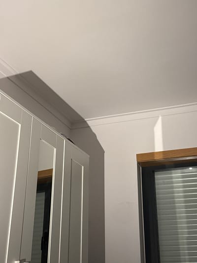 The image depicts an interior corner of a room, likely in Bragança, Portugal. The upper portion of the scene features a white ceiling with decorative crown molding along the junction with the walls. The walls are light-colored, possibly white or light grey. In the lower left quadrant, the upper section of a light-colored cabinet or wardrobe with recessed panel doors is visible. The cabinet's surface reflects portions of the room, including a dark-framed window. Above the cabinet, a prominent, elongated shadow is cast onto the ceiling and upper wall, extending from the cabinet's edge diagonally towards the upper right.
In the lower right quadrant, a window with a dark frame and horizontal blinds is present. A wooden lintel or trim piece is positioned directly above the window frame. A vertical band of bright light, potentially from another light source or a reflection, is visible on the wall to the left of the window and below the crown molding. No individuals, discernible actions, or direct interactions are visible in the static indoor environment.