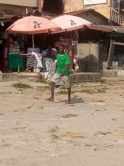 This image depicts an outdoor street scene in Badore, Nigeria, under daylight conditions.
In the foreground, an unpaved, dusty ground with uneven terrain and scattered debris, including a small red object and yellow fabric pieces, extends across the frame. A young adult male, dark complexion, is positioned slightly right of center, mid-stride, facing partially left. He wears a short-sleeved green shirt with a subtle pattern, light-colored (possibly khaki or grey) shorts, and brown flip-flops. A dark chain or strap is visible around his neck, and he holds a small, light-colored object in his right hand.
In the midground, a raised concrete curb separates the unpaved area from a retail stall or shop operating beneath two large, light-pink Coca-Cola branded market umbrellas. To the left, a dark-skinned individual in a blue shirt is partially visible behind a green counter, with various bottles and containers displayed. A Coca-Cola cooler stands upright on the far left. To the right, two dark-skinned individuals are seated; one wears a blue patterned shirt and faces left. A white sign reading "Deposit, Transfer, Withdrawal" is prominent, indicative of a financial service point. Another sign, partially visible, reads "CAMPENS" or similar. Various merchandise, including small packaged items, hangs from a display near the right umbrella.
The background consists of older, light-colored buildings with corrugated metal roofs. A dark arched doorway is visible in the central background building. A metal railing or staircase is present on the right side of a background structure. The overall environment suggests an urban or semi-urban setting with established, somewhat weathered infrastructure.