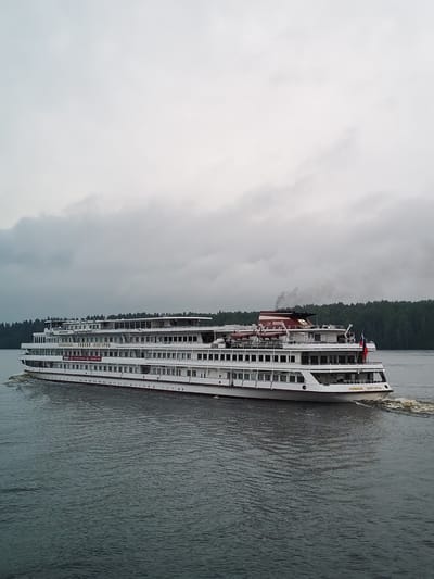 A large white passenger ship sails on a body of water under a cloudy sky. The ship has multiple decks with many windows. The name "НИЖНИЙ НОВГОРОД" is visible in gold lettering on the side of the ship. A red and white smokestack emits dark smoke. A Russian flag is visible on the stern of the ship. The water is dark and choppy, with a wake trailing behind the vessel. Dense green trees line the distant shore. The scene appears to be in the city of Khoroshevo, Russia. There are no people visible on the deck of the ship.