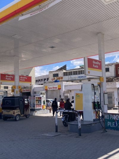 The image shows a Shell gas station, likely in Quetta, Pakistan. It is a daytime shot with moderate sunlight. There are four gas pumps visible under the station's canopy. A "Tyre Shop" is visible behind the station. Several people are present, some walking and some near motor vehicles. A three-wheeled auto-rickshaw is near one of the pumps. A motorcycle is parked near another. The overall condition of the facilities appears somewhat worn. A sign reads "NO PAK." There is also "Car Wash" sign posted.