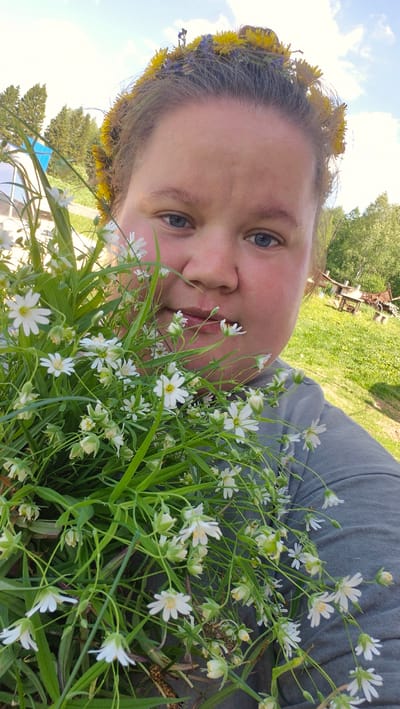 A medium shot depicts a person's face close to the camera, with a flower crown on their head and holding a bouquet of small white flowers. The person has fair skin, blue eyes, and is looking at the camera. The flower crown is made of yellow and blue flowers. The bouquet is composed of numerous small white flowers with green stems and leaves. The background includes grass, trees, and blue sky. The location is Дойная, Russia.