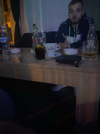A man in a Champion tracksuit sits at a table with alcoholic beverages and coffee. There is a glass of brown liquor, an ashtray with a lit cigarette, and a cup of Bianchi coffee. Bottles of liquor and water are also on the table. The location is Dobrovnitsa, Bulgaria.