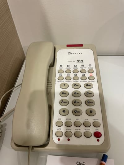 The image presents a beige Beetel hotel room telephone. The room number is designated as 312. The phone features buttons for front desk, housekeeping, room service, wake-up call, health centre, operator, concierge, laundry, restaurant, emergency, as well as standard number keys and function buttons like pause, volume, redial, and speaker. The telephone is situated on a glossy white surface with a light wood-grain surface visible in the bottom left, likely a tabletop or desk. There is a white piece of paper with writing on it beside the telephone, as well as a pen.
