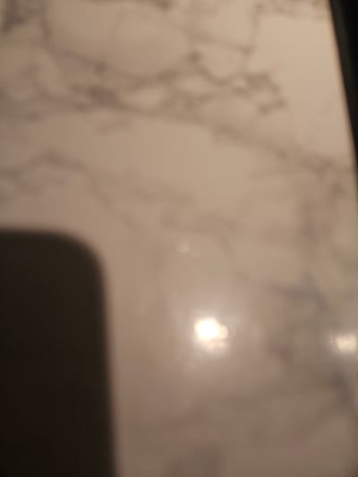 The image is a close-up, blurry, and out-of-focus shot of a reflective surface, possibly marble or a similar polished material. A dark, curved shadow occupies the lower left portion of the frame. Bright, bokeh-like reflections of light sources are visible in the mid-right and lower-right areas. The overall lighting is dim, with a focus on the reflective qualities of the surface. There is no discernible location in the city of Versailles, France, nor any identifiable people, subjects, or objects beyond the surface and the shadow.