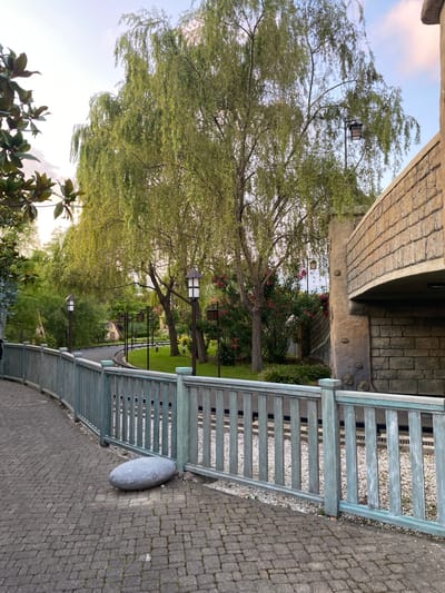 The image depicts an outdoor scene with a pathway and a fence. The pathway is paved with cobblestones and curves to the left. A light blue wooden fence with vertical slats runs along the right side of the pathway. Beyond the fence, there are train tracks and a grassy area. Several trees are visible, including a large willow tree with long, drooping branches in the center of the frame. There are also street lamps with lanterns along the pathway and in the background. To the right, a portion of a stone structure is visible, possibly a bridge or part of a building. The sky is visible at the top of the image, with soft clouds. In the foreground, near the fence, there is a large, grey, oval-shaped stone on the cobblestone path. The scene appears to be a park or an amusement park area. The image does not contain any people or specific landmarks that would indicate its location in the city of Sirius, Russia.