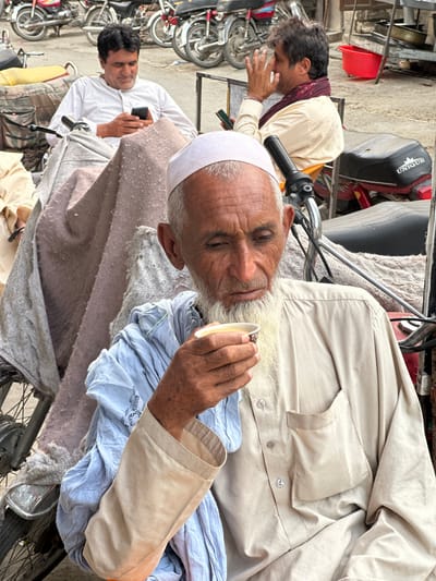 In Quetta City Tehsil, Pakistan, an outdoor scene unfolds with multiple individuals and motorcycles. In the foreground, an elderly man with a white beard and a light-colored shirt is holding a small cup to his lips, appearing to drink. He wears a light blue garment draped over his shoulders. Behind him, a middle-aged man in a white shirt and a maroon scarf is looking at his phone. Another man in a white shirt is also seated, and his phone is visible in his hand. Several motorcycles, including a red and black one with the branding "UNIQUE," are parked nearby, suggesting a roadside or market environment. The overall setting appears to be a public area, possibly a waiting spot or a commercial zone, with a slightly weathered and busy atmosphere.