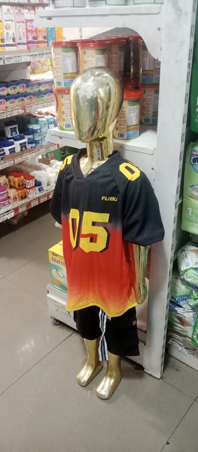 In Gwarinpa, Nigeria, a full-body mannequin with a metallic gold finish stands in an aisle of a retail store, possibly a pharmacy or supermarket. The mannequin is dressed in a black and red gradient jersey with yellow numbers "05" and the "FUBU" logo. It wears black shorts with white stripes. The mannequin is positioned in front of shelves stocked with various products. To the left, shelves display rows of small jars, likely cosmetics or toiletries, and larger containers of baby formula or similar products. To the right, shelves hold packs of diapers. The floor is tiled and appears to be a light grey color. The lighting is artificial, typical of an indoor retail environment.