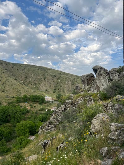 A mountainous landscape under a partly cloudy sky. The foreground features rocky terrain covered in vegetation. A hill slopes downward, dotted with trees, leading to a white building nestled among the greenery. In the upper left corner, power lines are visible against the sky. A unique rock formation resembling an animal head juts out from the top of the mountain.