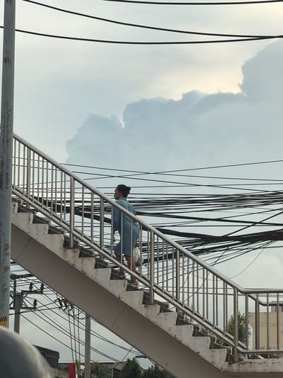 In an outdoor scene in Bangkok, Thailand, a woman with her hair in a bun, wearing a light blue shirt and denim shorts, ascends a concrete pedestrian overpass with a metal railing. She carries a translucent plastic bag in her right hand. The overpass stairs are worn, with visible concrete edges. Above and behind the overpass, a dense network of black electrical wires stretches across the sky, which is filled with a mix of light gray clouds and patches of bright sky. Buildings and palm trees are visible at the bottom of the image.
