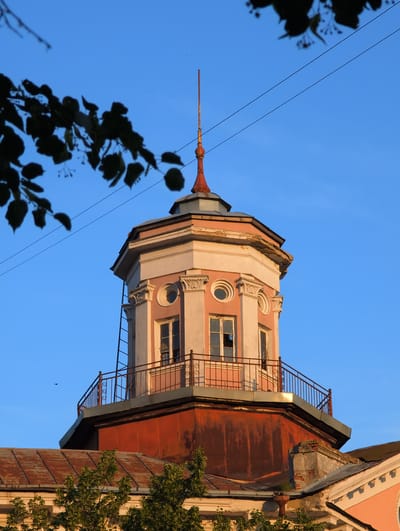 The image shows an old, ornate tower attached to a building, set against a clear blue sky. The tower is painted in pastel pink and cream colors, with decorative elements around its windows and at its peak. A rusty metal spire extends from the top of the tower.
The building itself appears weathered, with some areas of peeling paint and exposed brick, particularly on the rusted metal sections of the roof. There are windows visible on the tower, some of which are broken. A metal fire escape or balcony with decorative railings runs along the side of the building.
In the foreground, the dark silhouette of tree branches with leaves hangs into the frame, partially obscuring the view of the tower. Power lines are also visible crisscrossing the sky.
The lighting suggests it is either early morning or late afternoon, with the sun casting a warm glow on the building. The clear sky indicates fair weather. The architectural style hints at an older construction, possibly from the early 20th century. The scene is static, with no people or discernible activity taking place. The overall impression is one of historical architecture, showing signs of age and perhaps neglect, set against a peaceful, clear sky. There is no visible text in the image.