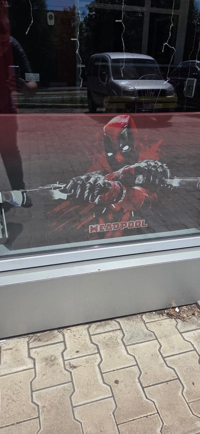 This image shows a storefront window with a large Deadpool graphic. The graphic depicts the character's masked face and hands holding what appears to be swords or weapons. The word "HEADPOOL" is visible in bold, white lettering below the character. Reflections in the window show a silver minivan and a person holding a phone, suggesting the photo was taken from the street. The exterior of the building has a grey metal frame around the window and a paved area with interlocked concrete blocks in front. The scene appears to be in an urban environment, likely on a street in Donetsk, Ukraine, given the context.