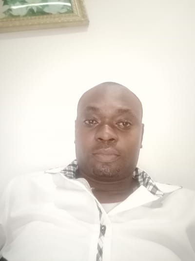 A head-and-shoulders shot depicts a dark-skinned, bald man wearing a white dress shirt with a checkered pattern on the inner collar. The background consists of a plain white wall with a framed picture hanging in the upper left corner. It is not possible to determine if the image was taken in Gwarinpa, Nigeria from this picture.