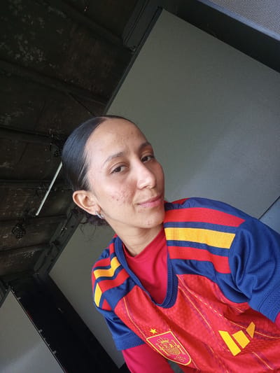 A medium shot captures a young woman from the chest up, looking directly at the camera. She has dark hair pulled back, revealing her forehead and ears. She wears a red shirt underneath a sporty blue, red, and yellow striped jersey, possibly a football uniform, with a crest on the left chest. Her facial features include brown eyes, a slightly upturned nose, and lips slightly parted in a gentle smile. Some blemishes are visible on her cheeks. She is wearing a small hoop earring in her left ear.
The background is divided, with the left side showing a dark, industrial-looking ceiling with exposed beams, lights, and electrical conduits. This suggests an indoor or covered outdoor space, possibly a stadium or venue. The right side of the background is a large, light gray, textured surface, resembling a screen or wall. The lighting appears to be a mix of natural and artificial sources, creating some shadows.
The overall impression is a casual, personal photograph, likely a selfie. There is no indication of a specific event or time of day beyond general daylight. The context of Porlamar, Venezuela, provides a geographical location, but no specific visual cues within the image confirm this. The woman's expression is relaxed and friendly. No visible text is present in the image.