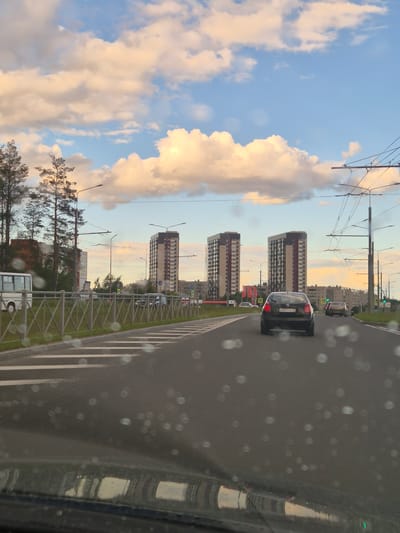 The image depicts a view from inside a vehicle, likely a car, looking out at a city street scene. The photo is taken during the day, with the sun low in the sky, casting a warm glow on the clouds, suggesting either early morning or late afternoon. The sky is a mix of blue and soft, illuminated clouds.

The primary subjects are modern, multi-story residential buildings with distinctive architectural features, characterized by their height and repeated window patterns. Several cars are visible on the road, including a dark-colored car in the foreground with its license plate partially obscured, showing Cyrillic characters and numbers: "P397TO". The road is asphalt with white lane markings, and a metal fence separates the road from a grassy verge. Streetlights with a distinctive design are also present.

In the background, more urban buildings and trees are visible, contributing to the city setting. The overall impression is one of a typical urban environment in Russia, specifically Petrozavodsk, given the context. The presence of water droplets on the surface from which the photo is taken (presumably the car's window) adds a subtle atmospheric detail, hinting at recent rain or dew. There is no clear indication of people actively engaged in an activity, but the presence of cars suggests everyday traffic and urban life. The scene is calm and ordinary, with no strong emotional cues beyond the pleasant lighting.