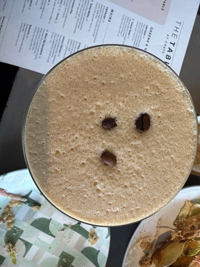 This is an overhead shot of an espresso martini in a glass, garnished with three coffee beans, alongside a menu from "The Table at Crate" and partially eaten plates. The location is likely a restaurant or bar, specifically "The Table at Crate", in Oak Brook, United States of America. The image suggests the end of a meal, with leftover food on the plates. The lighting appears to be natural or bright indoor lighting. The menu displays various food items, including salads, mains, sandwiches, and other dishes.