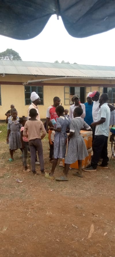 The scene is outside a building in Malava, Kenya. The building is yellow and has a corrugated metal roof. There are windows along the side of the building, and the windows are covered with window panes. The ground is dirt and there is dry grass on it. There are 12 people in the scene, including 5 adults and 7 children. The adults are wearing a variety of clothing, including shirts, pants, and hats. The children are wearing school uniforms. There are multiple objects in the scene, including an umbrella, a chair, and a water bottle. Some of the people are standing and some are sitting.  The adults are talking to each other, and some of the children are listening. There is a bicycle in the background. The scene is informal and the people appear to be engaged in casual conversation. The background is out of focus, but it appears to be a rural area. The people appear to be in the process of having a meeting. The scene takes place in front of a school building, possibly during a school event.