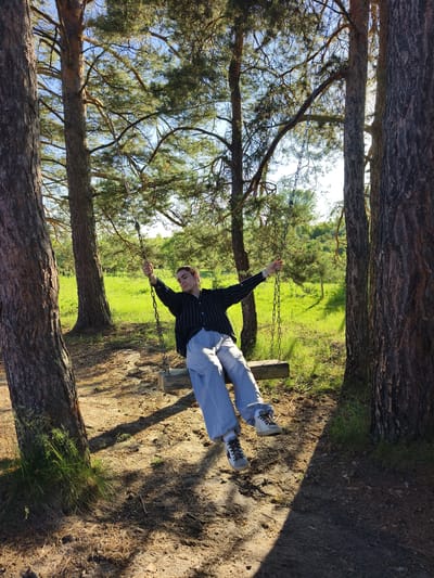 A young person is sitting on a wooden swing suspended from chains between two pine trees. The person is wearing a black and white striped shirt and light gray sweatpants. Their arms are outstretched, and their body is leaning back. The swing is in a natural outdoor setting with a grassy field and more trees in the background, under a clear blue sky. The ground is covered with pine needles and dirt, with sunlight casting shadows. The location is identified as Onokhino, Russia.