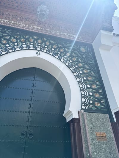 A low-angle shot captures a detail of the Bab Bhar gate in Tangier, Morocco. The scene focuses on the upper section of the gate, highlighting the ornate decorations surrounding a semi-circular arch above the door. The door itself is dark green and features vertical rows of rivets. Above the arch, intricate geometric patterns in black, white, and green tilework are visible. A plaque with Arabic script is affixed to a section above the tilework. A portion of a heavily carved wooden structure is seen on the left side of the image, possibly part of the gate's surrounding frame. The sunlight glares from the upper left.