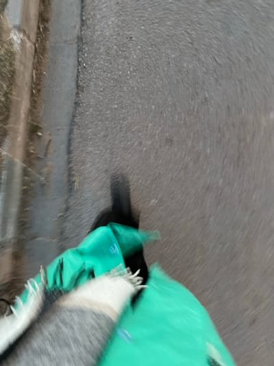 The image captures a motion-blurred, ground-level view of a scene featuring a bright turquoise or green object, possibly a bag or piece of fabric, on the right side. This object has visible folds and creases. Next to it, or partially draped over it, is a second textile object, likely a blanket or scarf, displaying prominent white and grey/dark stripes and distinct fringe or tassels along its edges.
A dark, elongated shadow is cast on the ground to the left of the green object, suggesting a light source, possibly the sun at a low angle or an artificial light. The ground itself is a grey, textured paved surface, consistent with asphalt or concrete, exhibiting significant motion blur that radiates outwards from a central point. Towards the bottom left, a faint, straight line indicates what appears to be the edge of a curb or a change in ground level, with hints of dry grass or dirt beyond it.
The heavy motion blur makes it difficult to discern a specific activity, but the presence of the objects on the ground suggests they are stationary or have been recently placed. There are no people visible. The time of day is not definitively clear, but the distinct shadow implies daylight, possibly dawn or dusk. There is a small, possibly circular, logo or symbol visible on the green object, but it is too indistinct to read any text. The location is contextualized as الغنادة, Tunisia.
