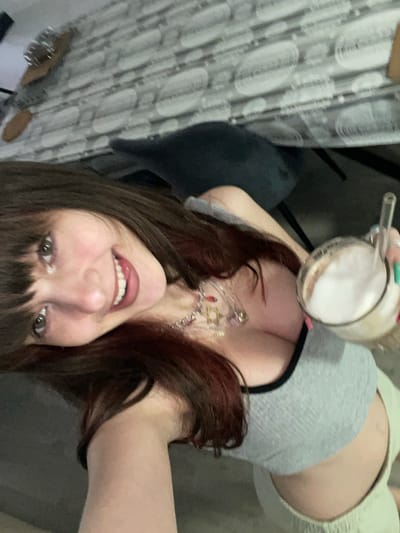 The image displays a female subject indoors, in what is described as the city of Raanana, Israel, captured in a tilted selfie orientation. The subject, positioned in the foreground, has long, dark brown hair with reddish undertones and bangs, and is depicted with a broad smile, showing white teeth. She is wearing a grey cropped tank top with black trim and light-colored bottoms. Around her neck is a layered necklace with multiple charms, including a prominent Star of David, a red heart, and additional small pendants. The subject's right hand holds a clear glass mug containing a frothy, light brown beverage with a clear glass straw. Her fingernails on the visible right hand display green and possibly pink nail polish. The background reveals an indoor setting, likely a dining or kitchen area, featuring a table covered with a grey and white patterned tablecloth. A dark grey upholstered chair is partially visible beneath the table, and additional objects, including a metal item and a wooden board, are present on the table surface in the upper left quadrant.