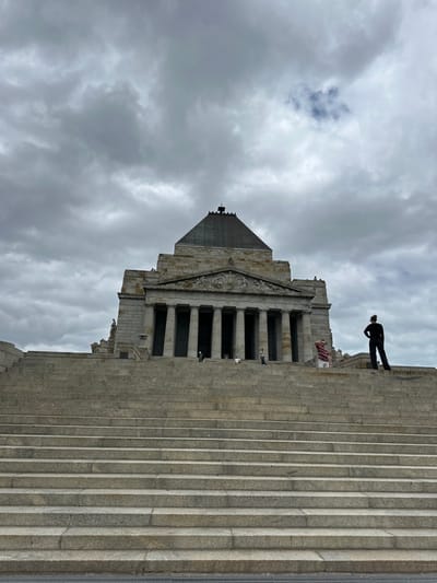 A symmetrical building with a pyramidal roof stands against a cloudy sky. The structure is constructed of gray stone and features a colonnaded facade. Wide steps lead up to the building's entrance. A person stands at the top of the stairs. The image is oriented in portrait mode, but rotated 90 degrees counter-clockwise.