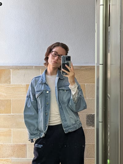 A young woman with short brown hair and glasses is taking a selfie in a hallway with tiled walls and what appears to be an elevator door to her right. She is wearing a light-colored long-sleeved shirt, an oversized denim jacket, and black sweatpants with blue lettering. The woman is holding a smartphone in her right hand, which is also reflected in a mirror. The lighting suggests it is daytime, and the environment appears clean and modern. There is no indication of the specific location within the city of Kiel, Germany.