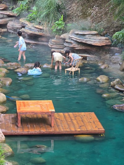 This image depicts a natural hot spring area in the city of Xiaoshan District, China, likely within a scenic tourist location. The water is a clear, vibrant turquoise color and appears to be shallow enough for people to stand in. Rocks of various sizes, some smooth and rounded, others layered and textured, line the edges of the spring and are submerged in the water. Lush green vegetation, including grasses and ferns, grows along the rocky banks and surrounding slopes. Steam rises gently from the water in several areas, indicating its warmth.
Four individuals are present in the scene. In the background, a young boy wearing a white t-shirt and shorts stands in the water, facing away from the camera. To his right, a child in a light-colored dress is bent over, appearing to interact with the water or the rocks. Further to the right, a young girl in a patterned dress is seated on a small, light-colored wooden chair placed in the water, also seemingly engaged with her surroundings. In the mid-ground, closer to the viewer, a young child in a blue dress is partially submerged in the water, facing away from the camera.
In the foreground, a wooden platform extends into the water, topped with a rustic, slatted wooden table. The platform and table are constructed from planks of warm-toned wood. The reflection of the table and platform is visible in the clear water below. The overall impression is one of a serene, natural environment designed for relaxation and enjoyment of the hot springs.