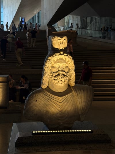 The image shows a large, stone bust of a man with a beard and elaborate headdress, illuminated by spotlights from below. The bust is situated in what appears to be a museum or public space with a large staircase in the background. Several people are visible on the stairs, some walking and some standing. The architecture features large, angular stone structures. The text on a plaque at the bottom of the image reads "Head of God Serapis". This suggests the sculpture is likely housed in a location that exhibits ancient Egyptian artifacts, possibly in or near Cairo, Egypt. The specific location of Kafrat Nassar is not visually confirmed by the image.