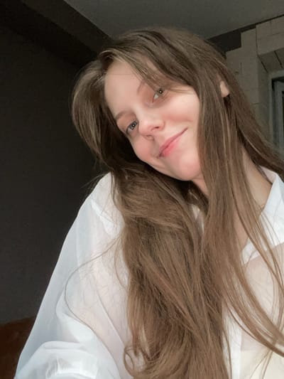 A young woman with long, brown hair is captured in a close-up, low-angle shot. She is wearing a white, possibly sheer, garment. Her face is partially obscured by her hair, but her light blue eyes are visible, looking towards the camera. She has a slight smile, with her lips parted. The background is dark and out of focus, with some blurry elements that might suggest an indoor setting, possibly with some brickwork visible in the upper left corner. The lighting appears to be dim, suggesting it might be nighttime or a poorly lit room. The overall mood of the image is intimate and relaxed. No text is visible in the image. The location context is Ufa, Russia.