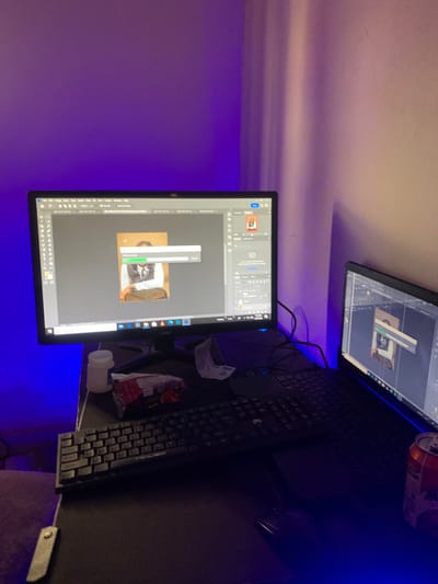 The image displays a dimly lit room with purple and blue ambient lighting. Two computer monitors are visible, one in the foreground and another slightly behind it. The foreground monitor shows a photo editing software interface, with an image of a person in a vest and shirt being edited. A progress bar is partially filled, suggesting an ongoing process. The background monitor displays a similar interface, also with an image being edited.
On the desk, a keyboard is positioned in front of the monitors. To the right of the keyboard, a soda can is precariously balanced on the edge of the desk. Various items are scattered on the desk, including what appears to be a small white container and a crumpled plastic bag. The overall setting appears to be a home office or workspace. The time of day is not discernible, and there are no weather cues. The image provides no specific location cues that would indicate Gwafan, Nigeria, beyond what is stated in the prompt. There is no visible text in the image that provides additional information.