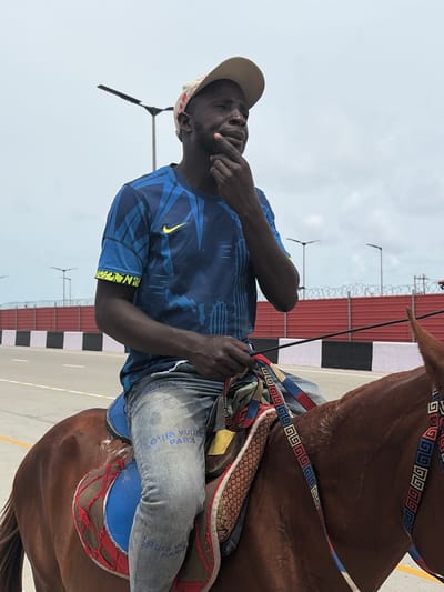A man wearing a tan baseball cap, blue shirt with a yellow Nike logo and blue patterned sleeves, and light blue jeans with "LOUIS VUITTON PARIS" printed on them, is sitting atop a brown horse. The horse has decorative straps and a saddle. The man is holding a black stick and has his hand near his mouth. In the background is a red fence, lampposts, and a light gray sky. The location appears to be an outdoor area in the city of Alaguntan, Nigeria.