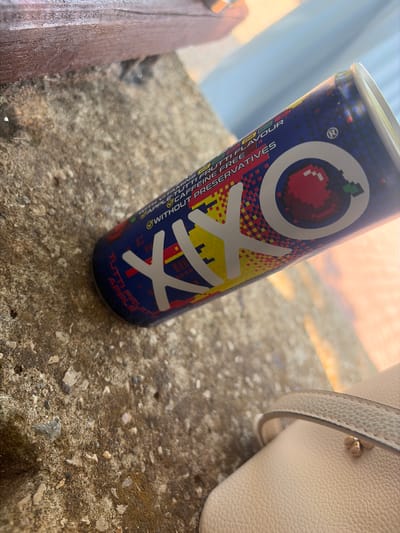 An outdoor shot, from a slightly low angle, shows a blue and yellow can of Xlxo energy drink resting on a rough, textured stone surface, possibly a step or wall. The can is positioned diagonally, with the top right fading out of focus and into a bright, hazy background suggesting sunlight. The can's label is partially visible, displaying the brand name "Xlxo" in large, stylized white letters and pixelated graphics. Text on the can indicates "WITH VITAMINS B6 B12 FRUTTI FLAVOUR CAFFEINE FREE WITHOUT PRESERVATIVES". A small, pixelated red apple graphic is visible on the can. The stone surface is gritty and has patches of dark and light brown, with small pebbles embedded. In the upper left corner, a blurred section of a wooden surface is visible. In the lower right, a portion of a light-colored handbag with a thin strap is in soft focus. The lighting suggests it is daytime with strong sunlight. The location of Vratsa, Bulgaria is not discernible from the visual elements of the image.
