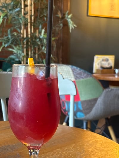 The image shows a close-up of a red beverage in a wine glass, positioned on a wooden table. A black straw extends into the glass, and a small piece of orange garnish is visible on the rim. In the background, a plant with green leaves is out of focus, and behind it, a wall displays two framed items: a yellow square frame on the left and a smaller, colorful framed picture on the right. Further in the background, a chair with a patchwork upholstery is visible. The lighting suggests it is daytime. The scene appears to be indoors, possibly a cafe or restaurant in Kragujevac, Serbia.