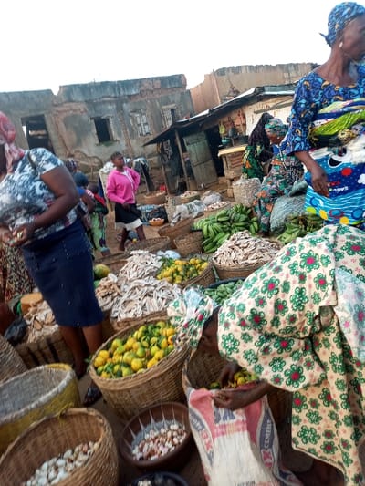 A bustling market scene unfolds with several women in patterned clothing attending to baskets overflowing with diverse produce. Fruits, dried fish, and other food items are neatly displayed in woven containers. The backdrop reveals aged buildings, makeshift stalls, and other people milling about, painting a vivid picture of a vibrant, everyday marketplace.