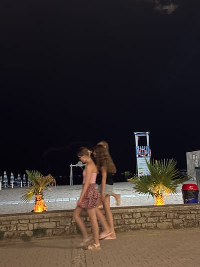 Three young people are walking along a paved walkway at night on a beach in Boreti, Montenegro. The sky is dark, with some clouds visible. On the beach, there are rows of closed beach umbrellas and a lifeguard tower with a life preserver. There are also two small palm trees illuminated with artificial light. A stone wall separates the walkway from the beach. One of the individuals is wearing a pink top and a patterned skirt, while another is wearing a black top and grey shorts. The third individual's clothing is not clearly visible. The overall scene suggests a calm evening on a beach resort.