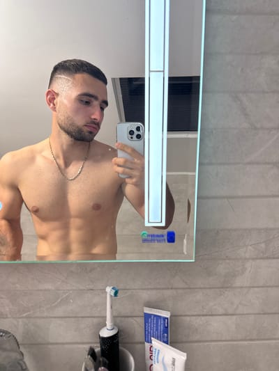 A forensic examination of the image reveals a young adult male, shirtless, with a muscular physique, short dark hair featuring a faded undercut and a visible curved line design on the left side, and a short beard. He wears a thin gold-colored chain necklace. The subject is taking a mirror selfie, holding a silver-colored smartphone with a triple-lens camera in his right hand. He is reflected in a wall-mounted mirror with integrated vertical LED lighting.

The setting is a bathroom, likely located in Gabrovo, Bulgaria. The walls are clad in large, horizontally arranged grey tiles, possibly with a marble or concrete texture. Below the mirror, a bathroom counter holds a white electric toothbrush with a blue and white brush head, standing in a white holder. Also visible are various hygiene products, including a partially visible tube with blue and white packaging, showing text such as "AQUA" and "ANTIPLAC," along with Cyrillic script and product numbers like "00140." Reflections in the mirror show a plain white ceiling, a window with blinds or shutters, and indistinct blue and white logos or text, one of which appears to be "Метелков." The overall lighting is a combination of natural and artificial light from the mirror's integrated LEDs.