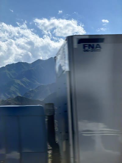 This media file depicts a view from a vehicle, likely a truck, as it travels. The perspective is from inside or on the roof of the truck's cab, looking towards the trailer and out into the landscape. The main subjects are the truck's trailer, a vast expanse of sky with cumulus clouds, and a rugged, mountainous terrain.
The truck's trailer is silver and partially visible, with the text "FNA" and some smaller lettering below it visible on its side. This suggests a commercial vehicle. The sky is a bright blue, indicative of daytime with fair weather, although the presence of substantial clouds suggests potential for varied weather.
The mountainous landscape in the background is lush and green, with deep valleys and steep slopes. The lighting suggests it is likely daytime, with sunlight casting shadows and highlighting the contours of the mountains. There's a sense of depth and scale to the scene, emphasizing the vastness of the natural environment.
The activity taking place is travel, with the truck moving through or alongside this scenic mountainous area. The image captures a moment during this journey. There are no people visible. The overall mood is one of motion and observation of a beautiful natural landscape from a utilitarian viewpoint, that of a vehicle in transit. The motion blur suggests the image was taken while the vehicle was moving.