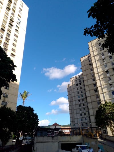 The image captures a view of a residential area in Caracas, Venezuela, likely the 23 de Enero sector, characterized by tall apartment buildings and lush greenery.
In the foreground, a white pickup truck is parked near a concrete underpass, with three individuals standing close to it. Two men are visible, one in a gray t-shirt and the other in a white t-shirt. A woman in a white top and dark pants is also present, holding a white bag. Graffiti adorns the concrete walls of the underpass.
Dominating the background are several high-rise apartment buildings. The building on the far left is a stark white, multi-story structure with numerous balconies. To its right, a series of beige apartment buildings are clustered together, their facades marked by rows of windows and balconies. A single palm tree stands between the white building and the beige ones. Further to the right, another large beige apartment building is visible. The sky above is a clear blue, punctuated by scattered white clouds.
The scene is set in a dense urban environment, with the apartment buildings suggesting a high population density typical of Caracas. The presence of trees indicates a balance between urban development and natural elements. The time of day appears to be daytime, with bright sunlight casting shadows, suggesting a clear weather.