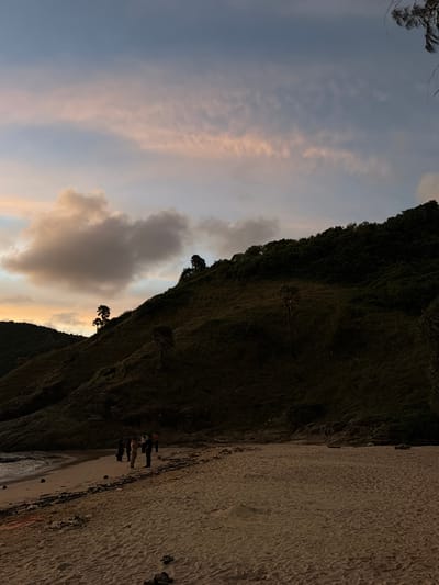 The image depicts a beach scene in Rawai, Thailand, at dusk. A group of approximately six individuals are visible on the sandy shore near the water's edge. They appear to be standing and interacting casually. The background features a dark, densely vegetated hillside that slopes down towards the beach. The sky above is a gradient of muted blues, grays, and pinks, with scattered clouds, indicating the setting sun. The sand is textured with footprints and a few scattered rocks and debris. The ocean is visible on the left side of the frame, with gentle waves lapping at the shore. A few silhouetted trees are present on the hillside, adding to the natural landscape.