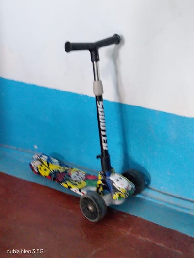 A three-wheeled scooter is leaning against a wall. The scooter has a black handle and a black stem with the word "SCOOTER" written vertically in white letters. The deck of the scooter is decorated with a colorful, graphic print featuring cartoon characters and abstract shapes in yellow, blue, red, and white. It has three black wheels. The background consists of a wall with a blue stripe running horizontally across it, separating it from a white upper section. The floor in front of the wall is a reddish-brown color. The lighting suggests it is daytime. The phrase "nubia Neo 3 5G" is visible in the bottom left corner of the image. The setting appears to be indoors, possibly in a home or storage area, in Angren, Uzbekistan. There are no people visible in the image, and the scooter is stationary.